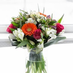 This beautifully designed bouquet embodies the refined artistry and local character of Upper Clapton florists. In a harmonious interplay of colour and texture, creamy beige and lively orange gerbera daisies radiate a cheerful warmth, ringed around plush, deeply toned magenta roses. Interspersed throughout, clusters of crisp white alstroemeria blossoms and delicate petite roses introduce a touch of grace and sophistication. Soft pink blooms and tiny white fillers accentuate the abundance, echoing the layered vibrancy of Springfield Park's springtime meadows. Robust, paddle-like aspidistra leaves are elegantly curved around the bouquet's edge, their glossy surfaces cradling the blooms and mirroring the fresh green of nearby towpaths. Narrower pointed foliage fills the spaces, adding dynamic structure and depth. The ensemble is displayed within a tall, slender glass vase, where the glowing green stems are clearly visible and remind one of nature's purity. Gentle, diffused daylight draws focus to the intricate design and lively palette, reminiscent of the uplifting atmosphere found in Upper Clapton's loved community corners. The bouquet conveys a sense of new beginnings, community celebration, and thoughtful care-ideal for brightening homes from Lea Bridge Road to every corner of E5. Each element reflects the florist's meticulous skill and the warm spirit known throughout the Upper Clapton neighbourhood.